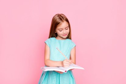 Lovely adorable charming little girl is looking down at the copybook in her hands and writing information there, she is wearing light blue dress, isolated on bright pink background, copyspace